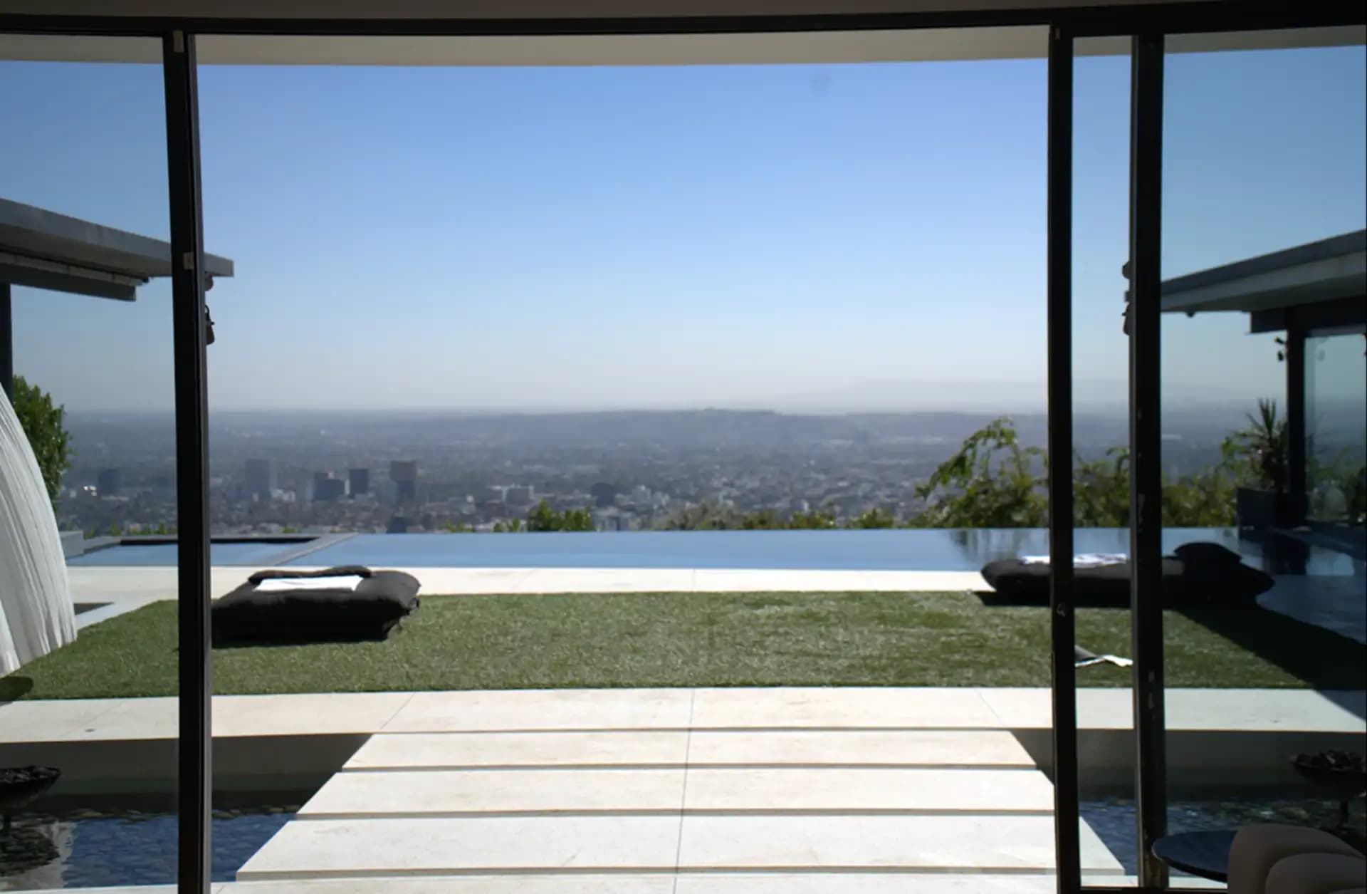View of a luxury home in Los Angeles with large glass doors overlooking the city from a modern infinity pool patio.