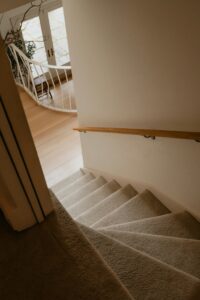 View of a clean carpeted staircase leading to a bright room with light wood flooring and natural light.