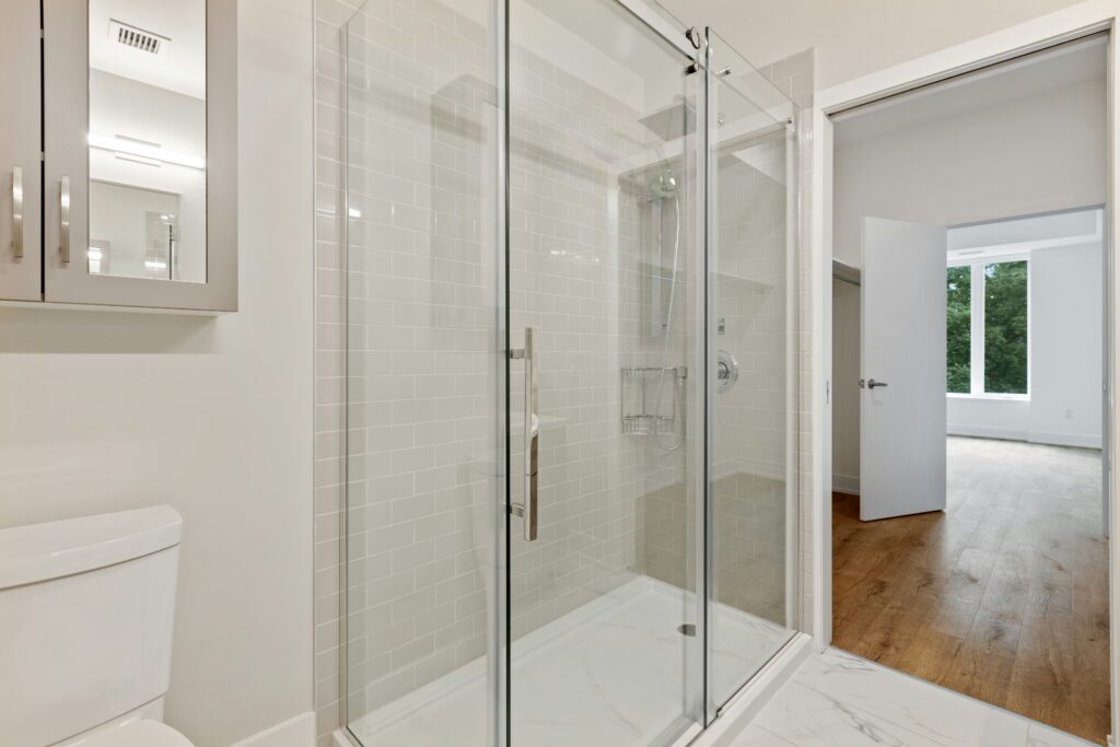 Modern bathroom with a clean glass shower enclosure, white tiles, and natural light from an adjacent room.