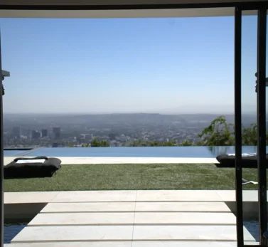 View of a luxury home in Los Angeles with large glass doors overlooking the city from a modern infinity pool patio.
